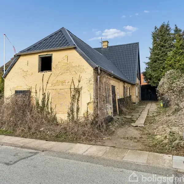 En gul murstensbygning står forladt. Facaden er overgroet med vegetation. En postkasse og et til salg-skilt med teksten "Hansen 24659133" ses ved fortovet. Husene omkring er beboede.
