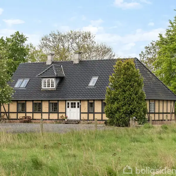 Et bindingsværkshus med sort tag står stille i en frodig have fyldt med grønne træer og græs. Det har flere vinduer og hoveddør midtfor.