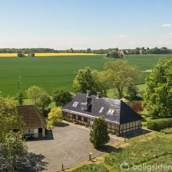 En bindingsværksejendom med sort tag er omgivet af gårdsplads og frodig have. Markerne strækker sig i baggrunden under en blå himmel med spredte skyer.