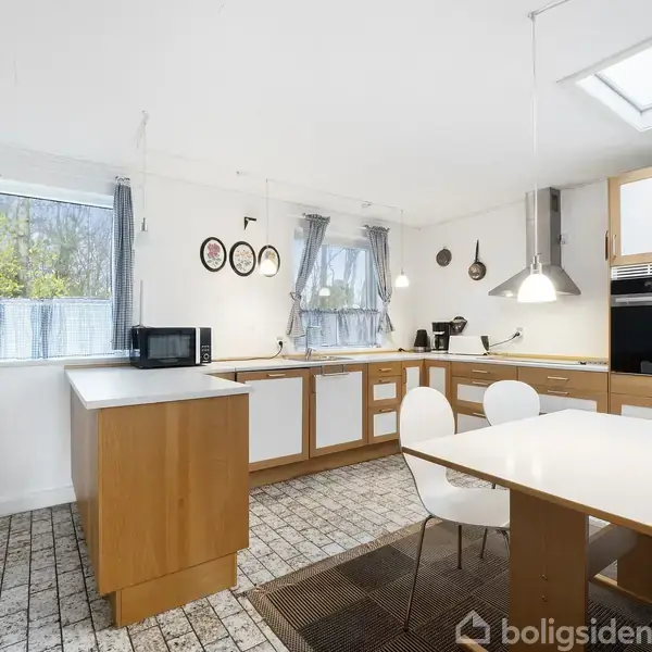 Køkken med træskabe, borde og moderne apparater, herunder mikrobølgeovn og stol ved bordet. Store vinduer med gardiner og vægophæng dekoreret med tallerkener. Flisegulv og loftsvindue.