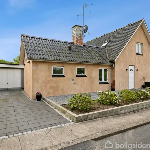 Hus med skråt tag og garage ved siden af, beliggende i et villakvarter. Foran er en indkørsel med fliser og en lille have med buske.