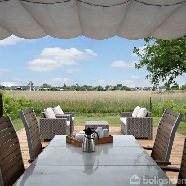 Havebord med ståltermokande står på en terrasse med udsigt over en mark. Træmøbler omringer bordet, og en pergola dækker pladsen. Træer og hække omkranser området.