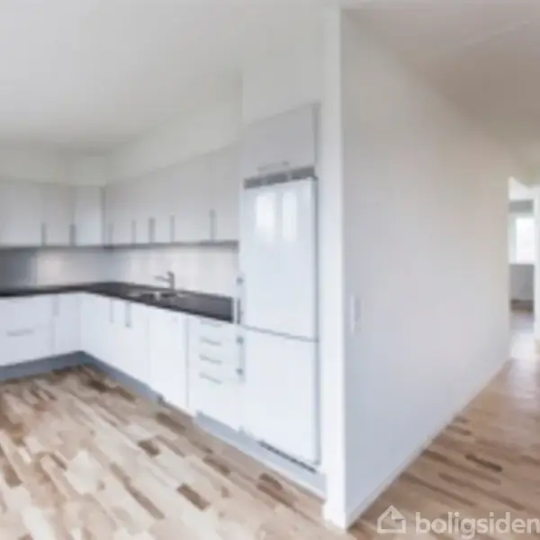 Køkken med hvide skabe og sorte bordplader, indeholder ovn og køleskab. Lys trægulv dominerer rummet, der åbner op mod en gang. Indtrykket er lyst og moderne.