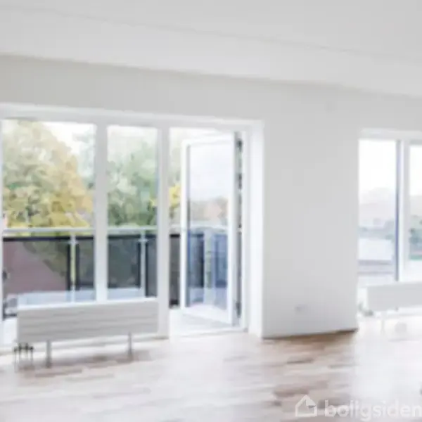 En lys stue med store vinduer, åben altandør med udsigt til grønne træer udenfor. Rummet har trægulv og hvide vægge, og der står en radiator under vinduet.