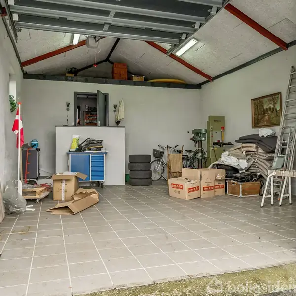 Garage med åbent dørparti indeholder stabile bananer-kartoner, cykler, dæk og en stige mod muren; opbevaringshylder fyldt med forskellige værktøjer og materialer. En lille dansk flag står oprejst.