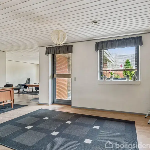 Stue med trægulv og loft, rektangulært tæppe på gulvet, skrivebord og kontorstol. Vindue med udsigt til have og entrédør. Hvidt træloft med rund lampe.