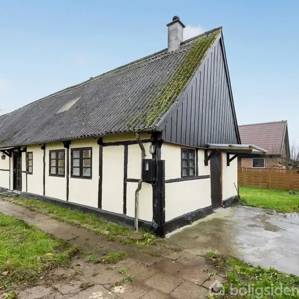 Bindingsværkshus med sort træværk og hvide vægge står på fugtig grund. Omkringliggende haver og træhegn skaber en landlig atmosfære under en lys himmel.