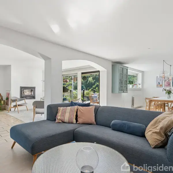 En blå sofa står i en moderne stue, der åbner ud mod en lys spisestue med trægulv. I baggrunden er der store vinduer og grønne planter.