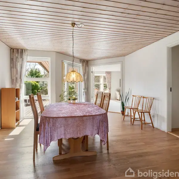 Et spisebord med lyserødt dug står i midten af en lys spisestue. Der hænger en lampe over bordet. I baggrunden er der store vinduer og døre, der leder udenfor.