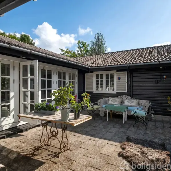 Gårdhave med bord og planter, omgivet af sorte trævægge og franske døre. En hvid bænk med puder står på fliserne, mens blå himmel og skyer ses i baggrunden.