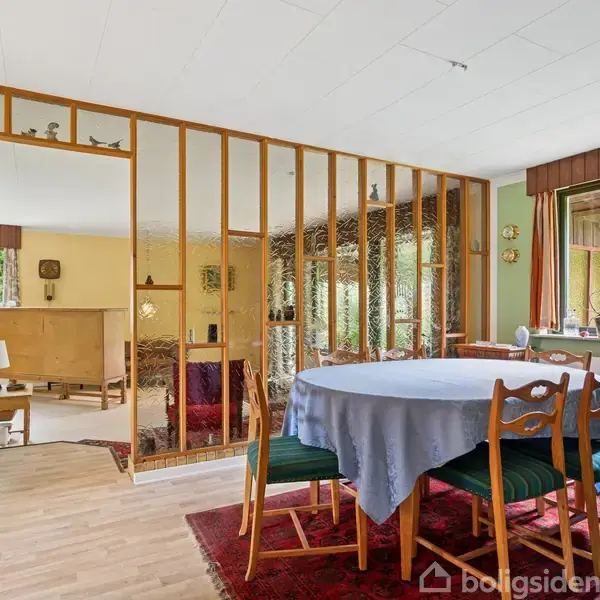Et rundt bord med blå dug og træstole står i en spisestue med lyst trægulv. Skillevæg af trælameller adskiller rummet fra en stue med sofaer. Store vinduer lader naturligt lys komme ind.