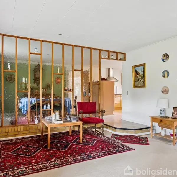 Rød stol og sofabord står på et mønstret tæppe i en stue. En trævæg med glas adskiller rummet fra spisestuen, hvor et dækket bord står i baggrunden. Malerier dekorerer væggen.