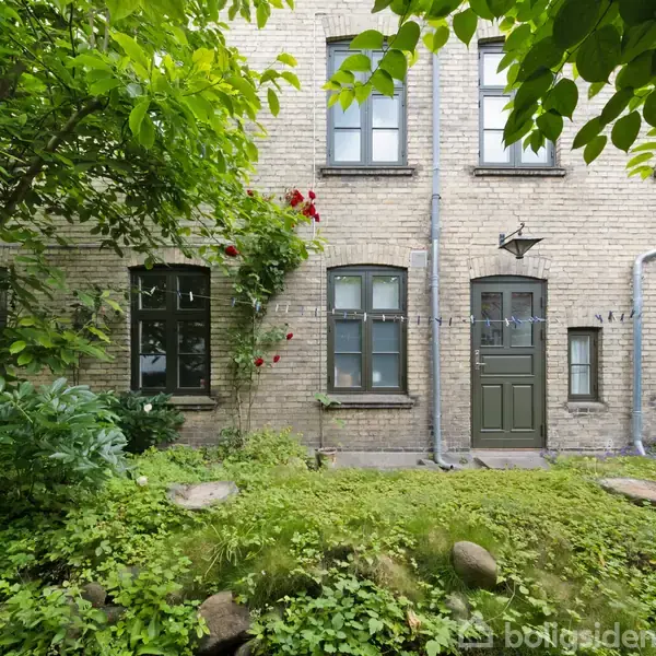 En murstensbygning med grøn hoveddør, omgivet af frodige planter og buske i en lille have, med røde roser ved siden af vinduerne og sten spredt på græsset.