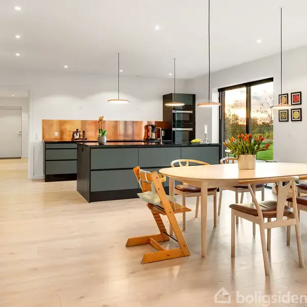 Et moderne køkken med spisestue. Bordet er omgivet af forskellige stole, tæt ved køkkenøen. Store vinduer giver udsigt til en grøn have. Lyse trægulve skaber et rum med varm atmosfære.