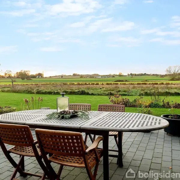 Et havebord med fire stole står på en flisebelagt terrasse. Bordet er dekoreret med en lanterne og planter. I baggrunden ses en stor, grøn græsplæne og åbne marklandskaber.