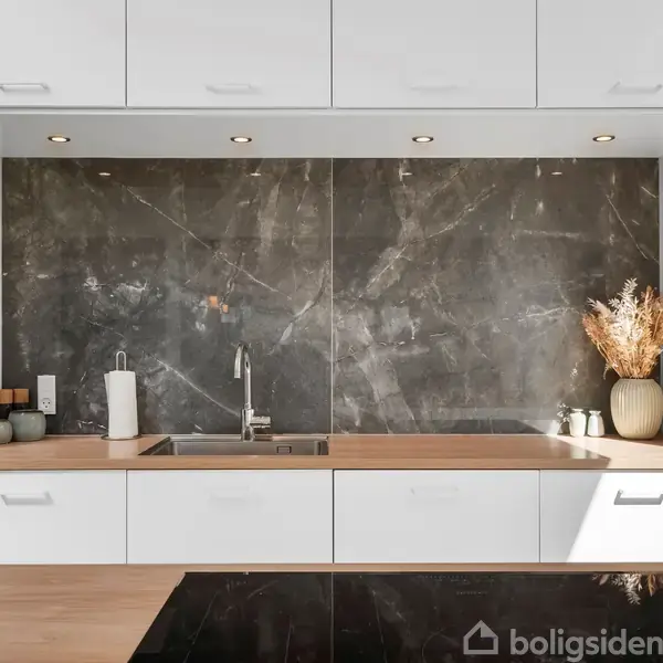 Et moderne køkken med en indbygget håndvask og hane i en træbordplade. En mørk marmor-bagvæg dekorerer rummet. På bordet står tørrede blomster i en vase og diverse køkkenartikler.