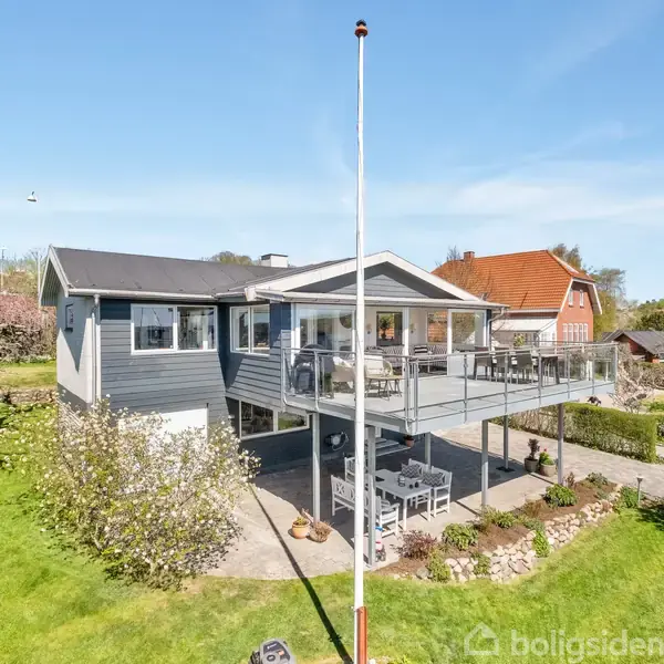 Et moderne hus med en stor altan og glaspartier. Det står på en velplejet græsplæne med blomstrende træer og omgivet af andre huse i et solrigt villakvarter.
