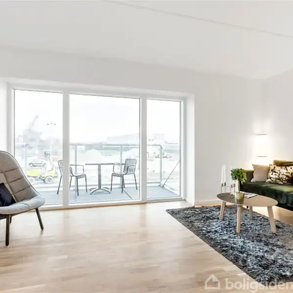 Stue med grøn sofa, bord og stol. En stor glasdør åbner til en altan med stole og bord. Lyse trægulve og en hvid væg skaber et moderne udtryk.