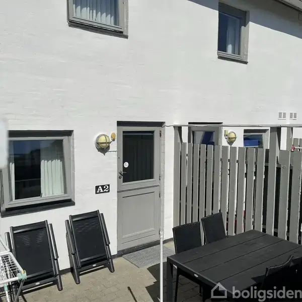 Dør med skiltet "A2" står ved siden af et vindue og en terrasse med sorte stole og bord. Sollys skinner på hvide vægge, og en grå træhegn afgrænser området.