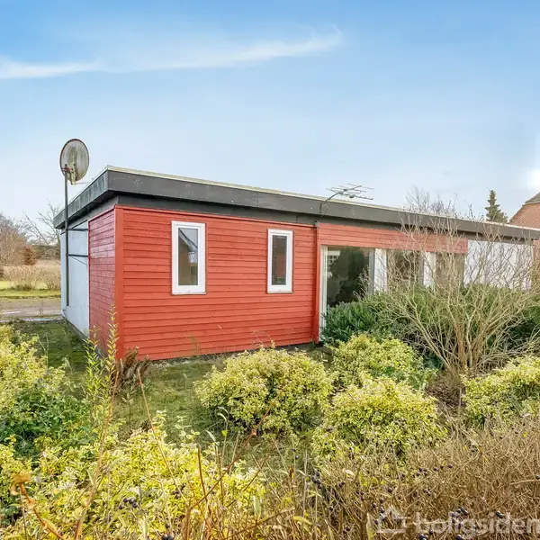 Rødt træhus med fladt tag står stille omgivet af buske og græs. Satelitantenne ses på taget under en blå himmel.