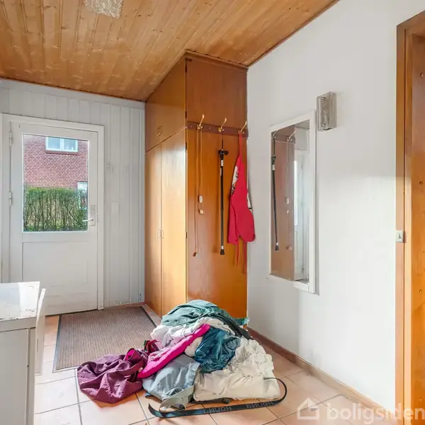 En bunke tøj ligger på gulvet foran et garderobeskab, som har tøj hængende på kroge. Der er en dør med glasrude mod venstre og en åbning til et rum med bogreoler til højre.