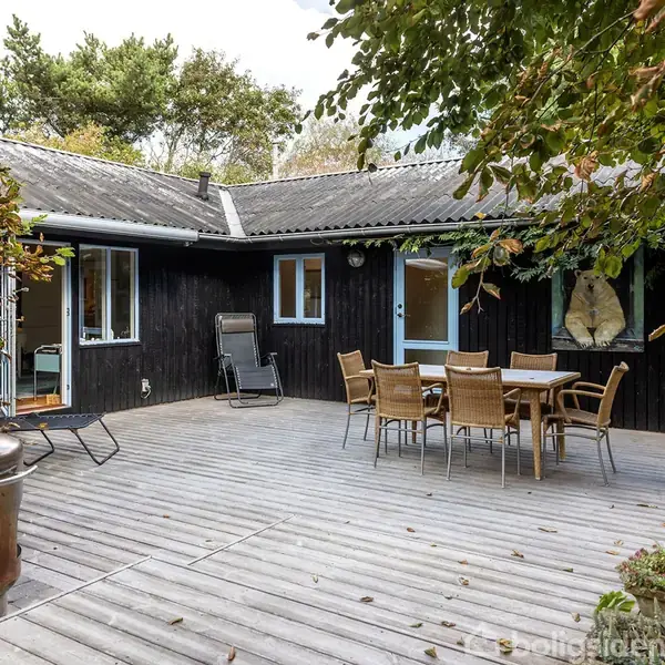 Sort hus med terrasse, hvor en kobberfarvet udendørsovn står. Et bord med stole er placeret under et træ. I baggrunden ses åbne døre, og omgivelserne er grønne.