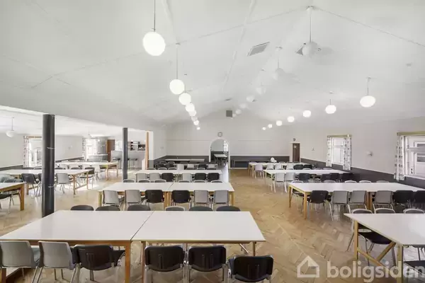 Et stort rum med mange rektangulære borde og stole arrangeret i rækker, oplyst af runde loftslamper. Trægulve og lyse vægge skaber en rummelig og neutral atmosfære.