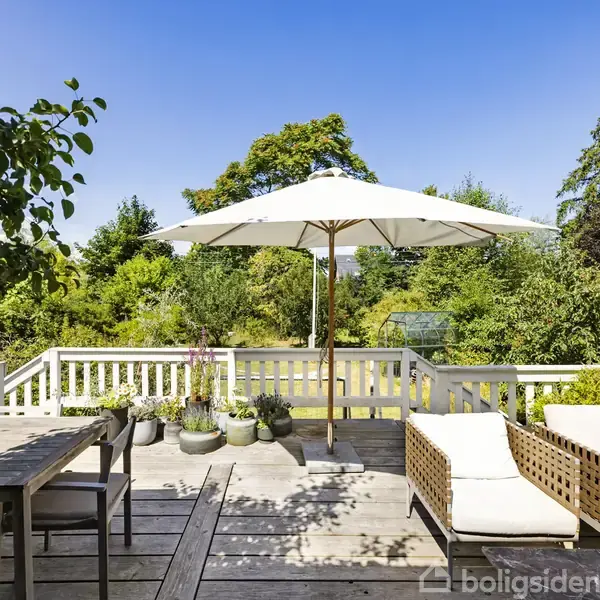 En terrasse med træmøbler og liggestole under en hvid parasol. Omgivet af grønne planter og træer, skaber dette et fredeligt udemiljø på en solrig dag.