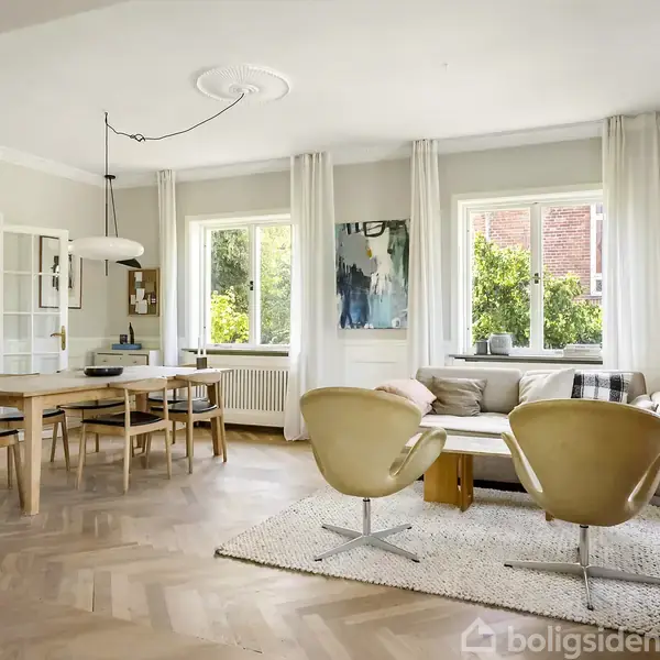 Spisestue med træbord og stole til venstre, og stueområde med sofa og lænestole til højre. Store vinduer med lyse gardiner slipper naturligt lys ind.