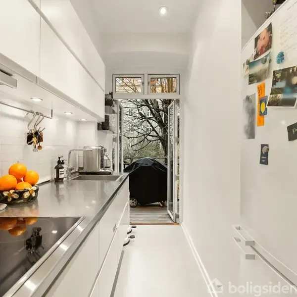 En smal køkken med hvide skabe og en sort bordplade, dekoreret med appelsiner i en skål. Et åbent vindue fører ud til en altan med udsigt til træer.