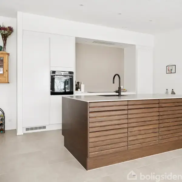 Køkkenø med træfront og sort vandhane står i lyst, moderne køkken. Omgivet af hvide skabe, integreret ovn, og pyntet med planter og en vinreol.