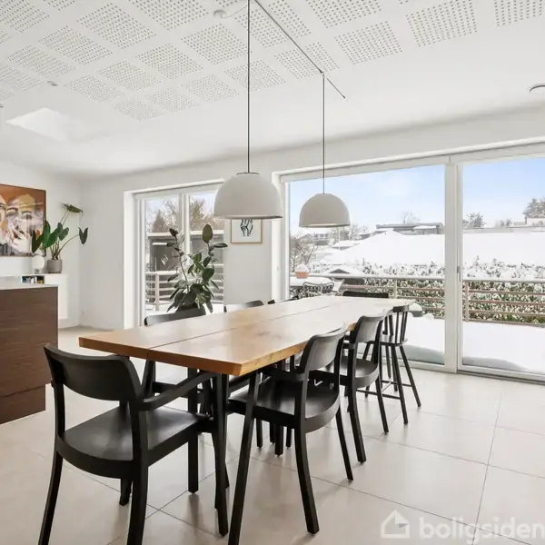 Et moderne spisebord med sorte stole står i et lyst køkkenområde. Store vinduer i baggrunden viser et snedækket landskab udenfor. To lamper hænger over bordet.