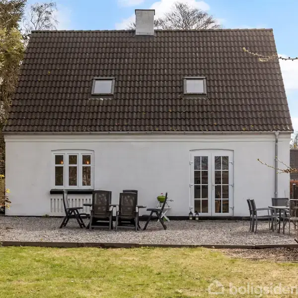 Huset er hvidt med et mørkt tegltag og to tagvinduer. I forgrunden er der en terrasse med havemøbler omkranset af græsplæne og buske.