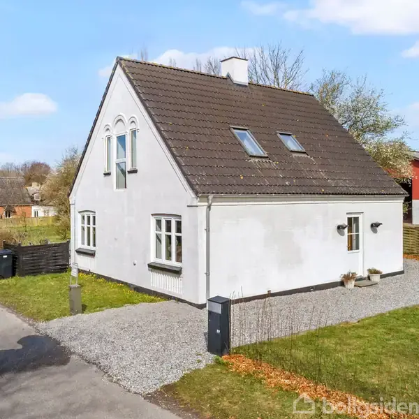 Et hvidt hus med mørkt tag står i et landskabeligt villakvarter. Huset har flere vinduer og en grusindkørsel. Omkringliggende træer og naboer kompletterer nabolaget.