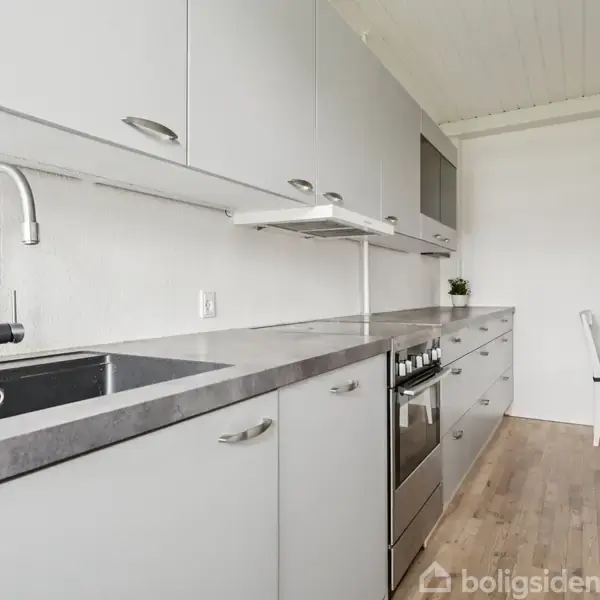 Et moderne køkken med lysegrå skabe og en mørk bordplade strækker sig langs væggen. En vask og kogeplade er synlige. En lille spiseplads med bord og stole i baggrunden.