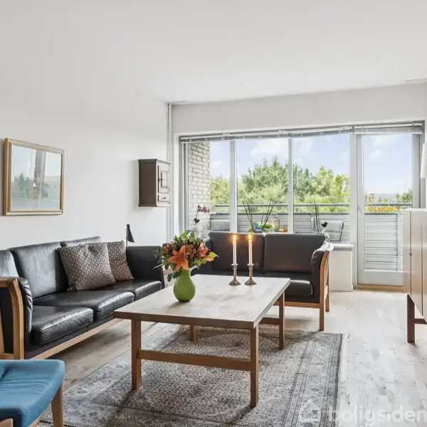 Sort lædersofa og lænestol omkranser et træbord med blomster og stearinlys. Rummet har en stor vinduesfront med udsigt til grønne omgivelser og minimalistiske møbler.