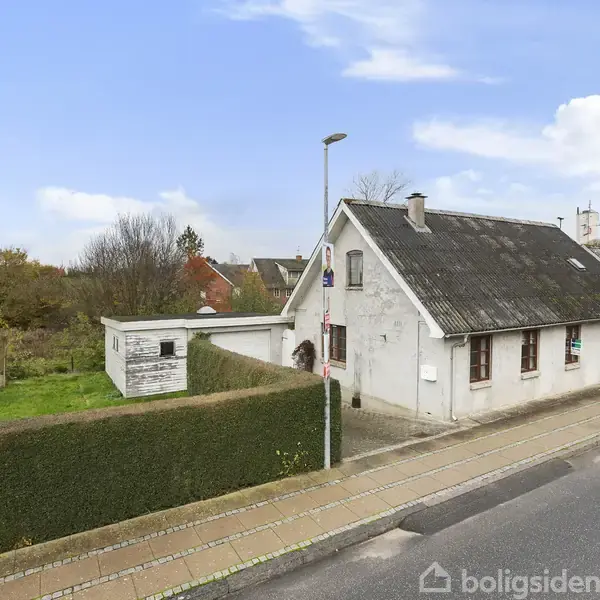 En hvid, ældre bygning med sort tag ligger ved en vej. Den er omkranset af grønne hække og græsplæne. Et mindre udhus er synligt i baghaven under en blå himmel.