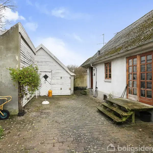 Et gammelt hus med mosbevokset tag og trædørtrin, står ved siden af en lille, hvid bygning i en gårdhave belagt med brosten, omkranset af hegn og et træ.