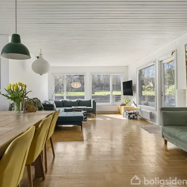 Et hus med store vinduer vender ud mod en flisebelagt terrasse. En orange bænk står til venstre. Omkring terrassen er der lave, grå stenmure med buske og træer i baggrunden.
