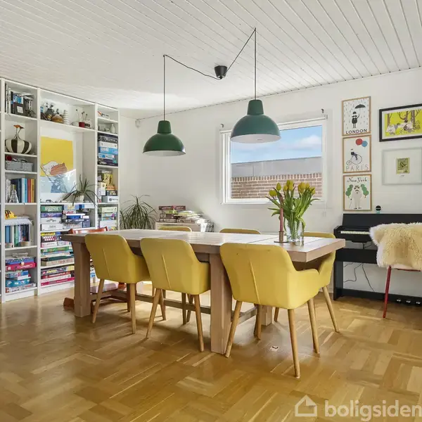 Langt bord med gule stole og en vase med blomster står i moderne stue. Kunstneriske plakater pryder væggene. Store vinduer giver udsigt til grønt haveområde udenfor.