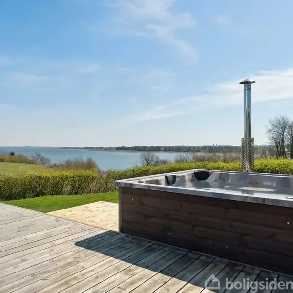 Udendørs spabad står stille på en træterrasse med udsigt over en sø og grønne bakker under en klar blå himmel. Skovtræer ses i baggrunden.