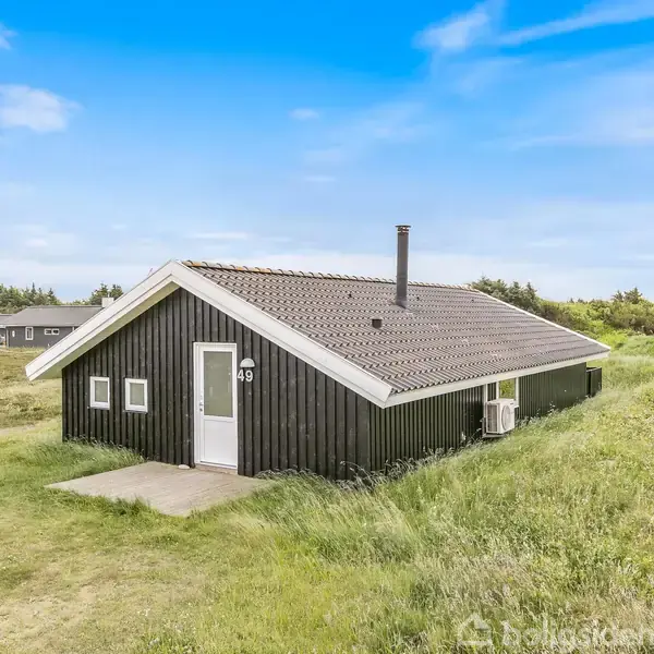 Et sort malet hus står stille omgivet af græs på en åben mark. Husets adresse er markeret med tallet "49" nær døren. I baggrunden ses flere små bygninger.