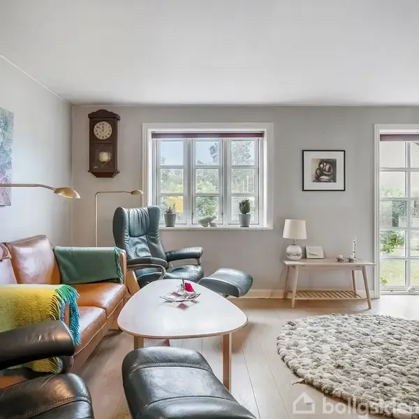 Stue med lænestole og sofa omkring et sofabord. Et stort vægur hænger på væggen. Rummet har lyse vægge, træmøbler, stort gulvtæppe og flere vinduer med udsigt til en have.