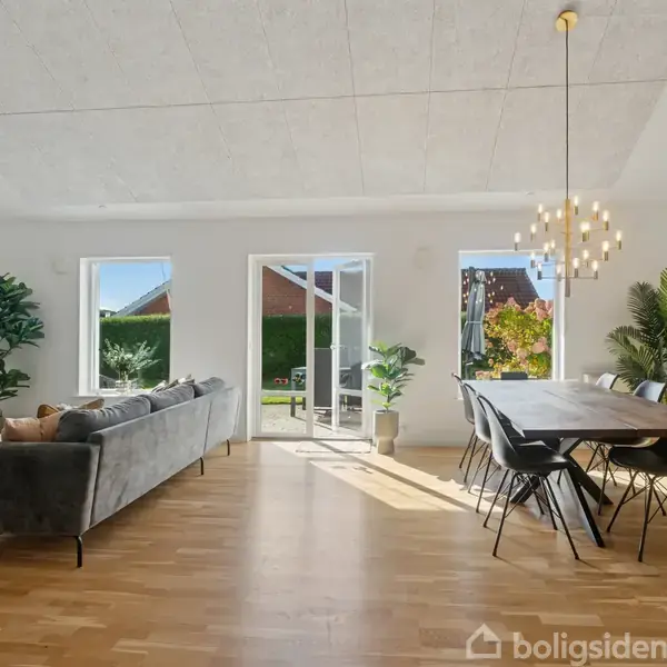 Sofa står i stue, ved siden af et spisebord, mens store vinduer lukker lys ind. Plantepynt og vægkunst skaber en moderne og lys atmosfære.