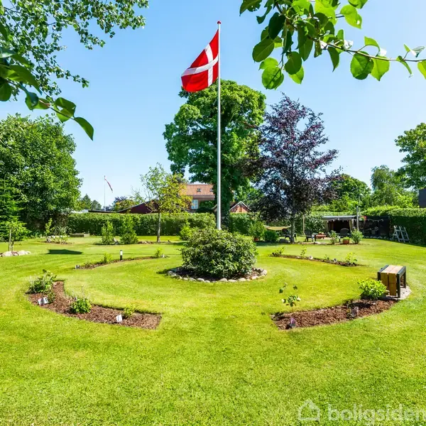 Flag på høj mast vajer i vinden, omgivet af velplejede græsplæner med blomsterbede, i en frodig have med høje træer og en privat bolig i baggrunden.