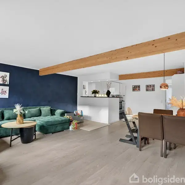 Grøn sofa i stuen står ved en blå væg med malerier. Sofabord i midten med dekoration. Til venstre legetøjskasse. Spisestue til højre med brunt bord og stole under loftlamper.