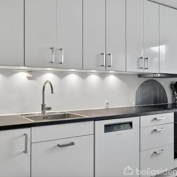 Køkken med hvide skabe og skuffer; armatur over vask; indbygget ovn til højre; oplyst arbejdsområde under overskabene; dæmpet, moderne stil.