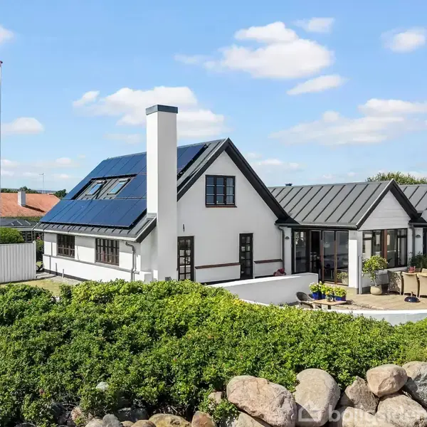 Et hvidt hus med sort tag har en flagstang med Dannebrog vajende. En terrasse med borde og stole omkranser huset. Omgivet af grønt buskads og blå himmel med skyer.