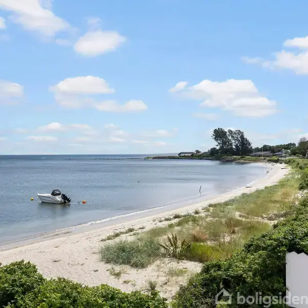 Båd ligger for anker ved en sandstrand med græs i forgrunden. Kystlinjen strækker sig mod horisonten, flankeret af et grønt område og få bygninger til højre.