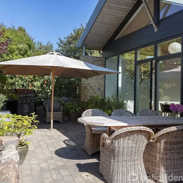 Havebord med kurvestole står under en parasol. Terrasse med brosten omgivet af grønne planter og træer. En grill er placeret til venstre ved siden af huset med store vinduer.